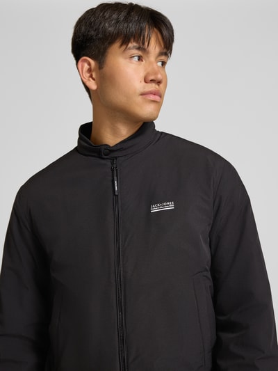 Männermodel trägt zwarte bomberjack met opstaande kraag, logoprint en ritssluiting, Model 'BROOKLYN' van JACK & JONES, regular fit, Nahaufnahme Oberkörper. Ruglengte maat M: 67 cm, schouderbreedte: 49 cm, borstwijdte: 59 cm, mouwlengte: 66 cm.