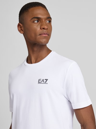 Männermodel trägt T-Shirt mit Label-Print von EA7 aus Baumwoll-Elasthan-Mix, Slim-Fit, Weiß, gerippter Rundhalsausschnitt, Nahaufnahme Oberkörper.