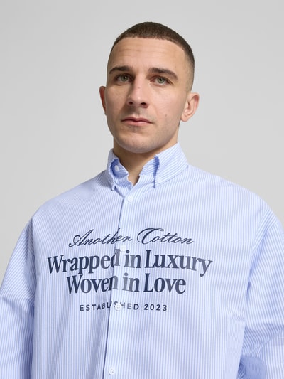 Męski model nosi Regular Fit Freizeithemd mit Statement-Print von ANOTHER COTTON LAB, koszula bawełniana w prążki, jasnoniebieska, krój regular fit, model 'Wrapped in Luxury', kołnierzyk button-down, zapięcie na guziki, nadruk na piersi, detal z logo, sportowe mankiety, długość pleców w rozmiarze M: 71 cm, szerokość w rozmiarze M: 53 cm, długość rękawa w rozmiarze M: 60 cm, zbliżenie na górną część torsu.