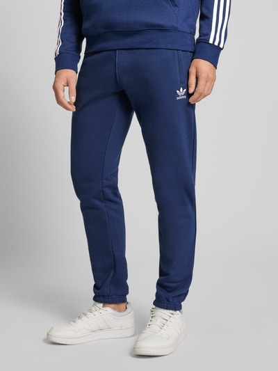 Mannenmodel draagt 'Sweatpants mit elastischem Bund' van adidas Originals in donkerblauw, katoenmix, losvallende pasvorm, label-stiksel, Franse steekzakken, elastische tailleband, binnenbeenlengte maat M 76 cm, voorkant geheel zichtbaar.