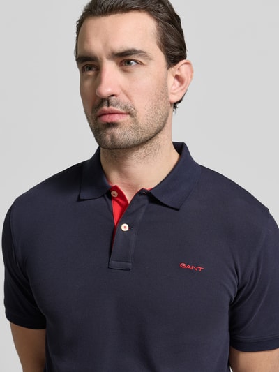 Mężczyzna nosi Regular Fit Poloshirt mit Logo-Stitching von Gant z elastycznej mieszanki bawełny, krój regular fit, polokołnierzyk, krótki rękaw, granatowy z czerwonym detalem, logo haftowane, widoczna faktura materiału, długość tyłu w rozmiarze M: 70 cm, zbliżenie na górną część ciała, przód