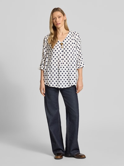 Frauenmodel trägt Blusenshirt mit Viskose-Anteil von Apricot, weiß mit schwarzen Punkten, V-Ausschnitt, Zierknöpfe, gerader Schnitt, 3/4-Arm, fließender Stoff, Ganzkörperansicht von vorne.