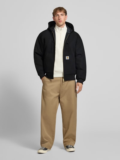 Männermodel trägt Jacke mit Label-Patch und Känguru-Tasche von Carhartt Work In Progress in Schwarz, kombiniert mit heller Hose, Ganzkörperansicht von vorne. Regular Fit, Kapuze mit Kordelzug, Reißverschluss, gerippte Abschlüsse, reine Baumwolle.