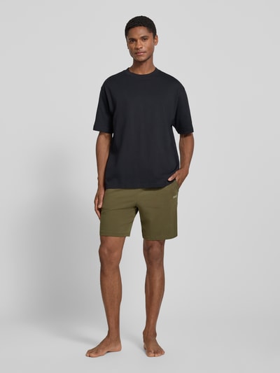 Männermodel trägt Regular Fit Sweatshorts aus Baumwoll-Mix Modell 'MIX&MATCH' von BOSS in Oliv, kombiniert mit schwarzem Shirt, Herren Sweatshorts, Ganzkörperansicht von vorne.