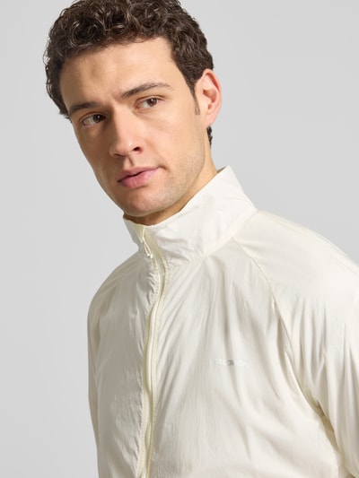 Männermodel trägt Jacke mit Stehkragen und Logo von Salomon in Creme, glatte Textur, Reißverschluss, Regular Fit, Nahaufnahme Oberkörper.