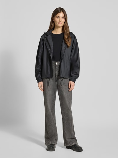Frauenmodel trägt Jacke mit Abdeckleiste von Rains, gerade Passform, schwarze Farbe, Kapuze mit Tunnelzug, Ganzkörperansicht von vorne. Rückenlänge bei Größe S 58 cm, Schulterbreite 43 cm, Brustweite 55 cm, Ärmellänge 63 cm, durchgehender Reißverschluss, elastische Ärmelabschlüsse.