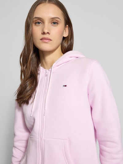 Vrouwenmodel draagt Regular Fit Sweatjacke aus Baumwoll-Mix van Tommy Jeans in lichtroze, unikleurig, met capuchon, kangoeroezak, logo-stiksel, sweatstof, katoenmix. Nahaufnahme bovenlichaam.