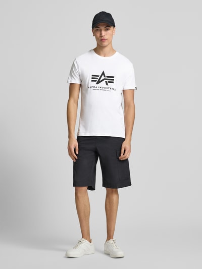 Männermodel trägt weißes Alpha Industries Herren T-Shirt mit Label-Detail, Rundhalsausschnitt, Gerader-Schnitt, Reine Baumwolle, Modell 'Basic', Rückenlänge 67 cm, Ganzkörperansicht von vorne.