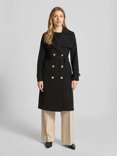 Modelka nosi czarny dwurzędowy trencz ‘Zweireihiger Trenchcoat mit Bindegürtel’ od MICHAEL Michael Kors, krój regular fit, kołnierz z połami, wiązany pasek, długość tyłu w rozmiarze S: 47 cm, szerokość ramion w rozmiarze S: 40 cm, szerokość klatki piersiowej w rozmiarze S: 50 cm, długość rękawów w rozmiarze S: 63 cm, jasne spodnie, prosty krój, gładka faktura, widok całej sylwetki z przodu.