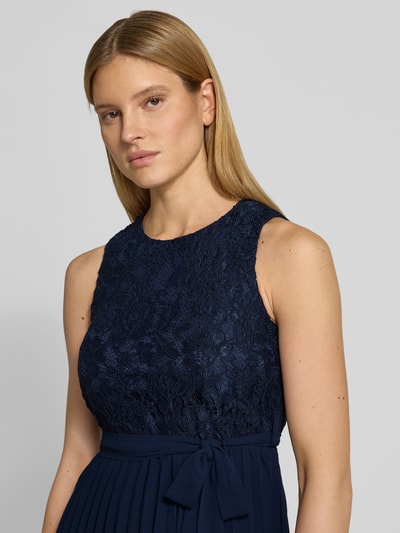 Kobiece model prezentuje Knielanges Kleid mit Spitze od Apricot, mieszanka materiałów z dodatkiem wiskozy, taliowany fason, długość do kolan, koronka, okrągły dekolt, wiązany pasek, spódnica z plisami, wycięcie w kształcie łezki z tyłu, szerokość w rozmiarze S: 39 cm, długość tyłu: 91 cm, ciemny granat, tekstura koronki, zbliżenie na górną część ciała, widok z przodu