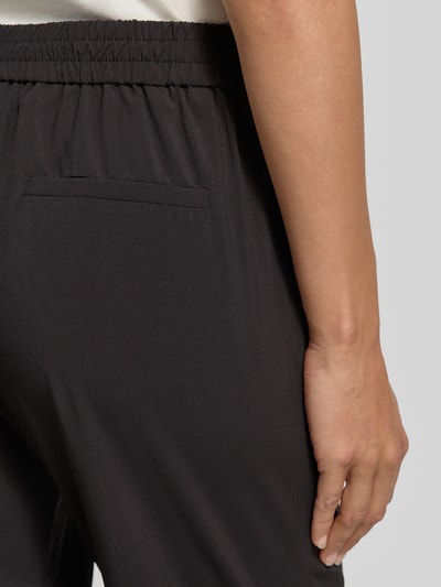 Vrouwenmodel draagt Hose in verkürzter Passform Modell 'Charlie' van Someday in zwart materiaalmix met stretch, elastische band, achterzak, detailopname heup. Context: dames broek met verkorte pasvorm, binnenbeenlengte 67 cm, bandwijdte maat 36 is 39 cm.
