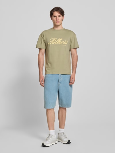 Mężczyzna model nosi t-shirt z nadrukiem z logo i okrągłym dekoltem BLKVIS, męski t-shirt z bawełny, krój regular fit, długość tyłu w rozmiarze M 68 cm, kolor khaki, jasnoniebieskie denimowe szorty, białe sneakersy, widok całej sylwetki z przodu.