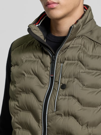 Męski model nosi Regular Fit Steppweste mit Kapuze im Kragen od Tom Tailor, oliwkowa, jednolita, krój regular fit, z label patch oraz odblaskowymi detalami, stójka z kapturem w kołnierzu, kieszenie boczne na zamek, zdjęcie w zbliżeniu na górną część torsu