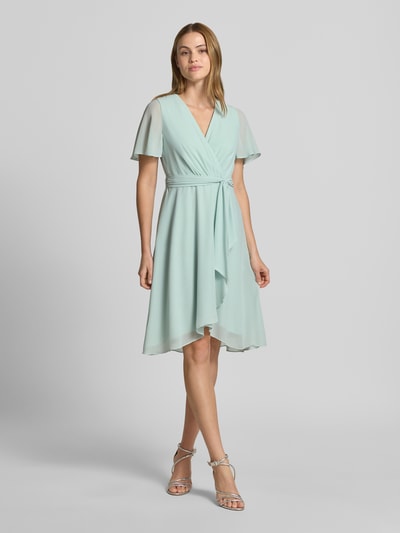 Frauenmodel trägt Knielanges Cocktailkleid in Wickel-Optik von Christian Berg Cocktail aus mintfarbenem Chiffon mit A-Linie, V-Ausschnitt, Bindegürtel, knielang, Ganzkörperansicht von vorne.