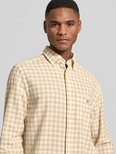 Męski model nosi Regular Fit Leinenhemd mit Baumwoll-Anteil Modell 'GINGHAM' od Gant, mieszanka lnu i bawełny, beżowo-biała krata, wyhaftowane logo, listwa guzikowa na całej długości, kołnierzyk typu button down, kieszeń na piersi, krój regular fit, z długim rękawem, zaokrąglony dół, długość tyłu w rozmiarze M: 66 cm, szerokość w rozmiarze M: 46 cm, długość rękawów w rozmiarze M: 64 cm, tekstura lekko miękka, widok: zbliżenie na górną część ciała.