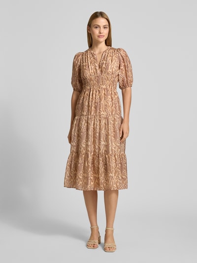 Frauenmodel trägt Midikleid aus Lyocell-Mix Modell 'Tanyssa' von Bash mit allover Paisley-Print, leicht glänzende Textur, taillierte Passform, Tunika-Kragen, elastische Ärmelabschlüsse, warme Beige- und Rosatöne, Ganzkörperansicht von vorne.