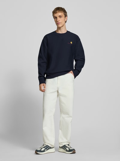 Männermodel trägt Regular Fit Sweatshirt mit Logo-Stitching von Carhartt Work In Progress in Dunkelblau, Baumwoll-Mix, Rundhalsausschnitt, weiße Hose, Ganzkörperansicht von vorne.