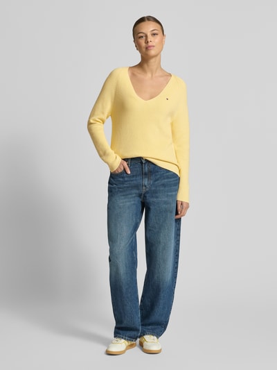 Kobiece model prezentuje Regular Fit Strickpullover aus reiner Baumwolle od Tommy Hilfiger w jednolitym żółtym kolorze, długi rękaw, V-dekolt, logo, struktura prążkowana, widok całej sylwetki z przodu.