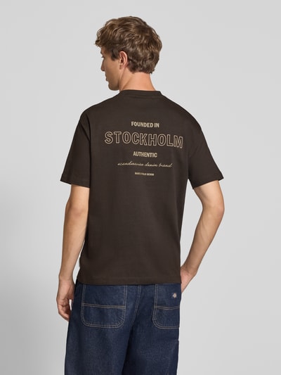 Männermodel trägt Relaxed Fit T-Shirt aus reiner Baumwolle von Marc O'Polo DENIM in Dunkelbraun, Rückseite mit Schriftzug, Ganzkörperansicht Rückansicht.