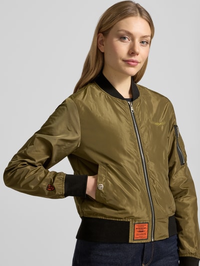 Frauenmodel trägt Bomberjacke mit Label-Patch und Reißverschlusstasche am Ärmel von Original Bombers, olivgrün, Glanz-Textur, schwarzer Stehkragen, elastische Abschlüsse, Logo-Stitching, Seitliche Eingrifftaschen mit Druckknopfverschluss, Nahaufnahme Oberkörper.