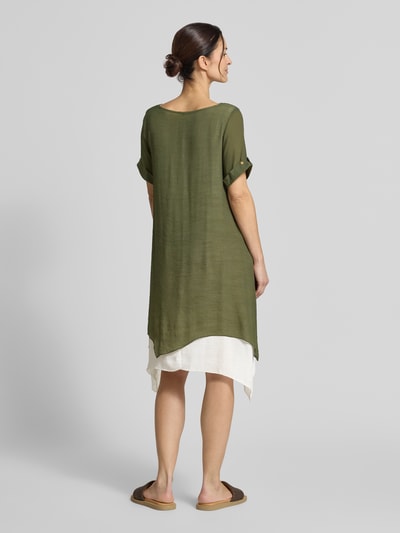Frauenmodel trägt knielanges Kleid aus Viskose-Mix von Apricot in Olive mit weißem Saum, asymmetrischer Form, Rückenansicht, Ganzkörperansicht.