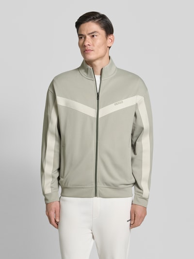 Męski model nosi Relaxed Fit Sweatjacke mit Baumwoll-Anteil Modell 'DARTI' od HUGO, jasnoszary kolor, zamek błyskawiczny, stebnowane detale, boczne kieszenie, jednolity wzór, chevron-tape na rękawach, długi rękaw, stójka, label-detail, prążkowane mankiety i dół, całkowity widok z przodu.