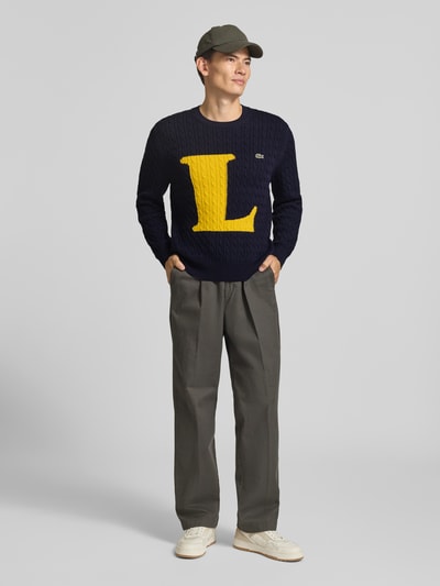 Männermodel trägt Regular Fit Strickpullover aus Baumwoll-Mix von Lacoste in Dunkelblau mit großem gelbem L, Ganzkörperansicht von vorne. Herren Strickjacke von Lacoste.