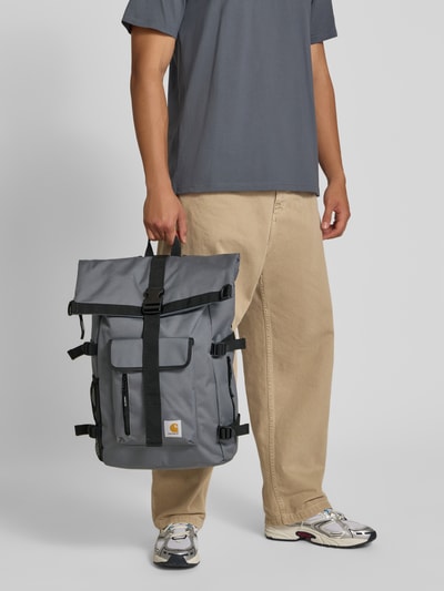 Männermodel trägt Rucksack mit Logo-Patch Modell 'PHILIS' von Carhartt Work In Progress in Grau mit schwarzem Gurt, Ganzkörperansicht von vorne. Hauptfach mit Überschlag, verstellbare Schulterriemen, Breite 52 cm, Länge 52 cm, Tiefe 14 cm, Riemenlänge 64 cm.