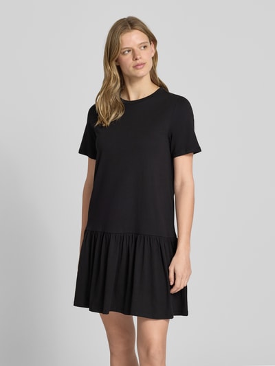 Modelka kobieca nosi Regular Fit T-Shirt-Kleid aus reiner Baumwolle Modell 'DISSO' od Pieces, czarna, krótki rękaw, falbaniasty dół, mini, gładka faktura, widok całej sylwetki z przodu.