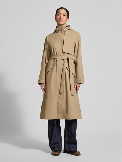 Frauenmodel trägt Regular Fit Mantel mit Stehkragen Modell 'Suva' von Rains, beige, taillierte Passform, Stoffband, Pattentaschen, Ganzkörperansicht von vorne. Rückenlänge bei Größe S: 113 cm, Schulterbreite: 42 cm, Brustweite: 51 cm, Ärmellänge: 62 cm.