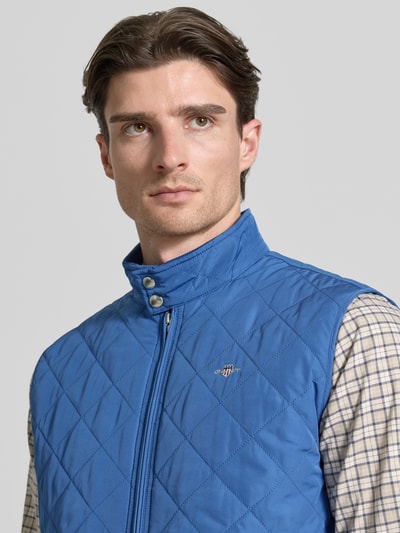 Männermodel trägt Regular Fit Weste mit Stehkragen in Stepp-Optik von Gant in Blau mit Logo-Stitching, Reißverschluss, Nahaufnahme Oberkörper. Rückenlänge bei Größe M: 69 cm, Schulterbreite: 45 cm, Brustweite: 57 cm.