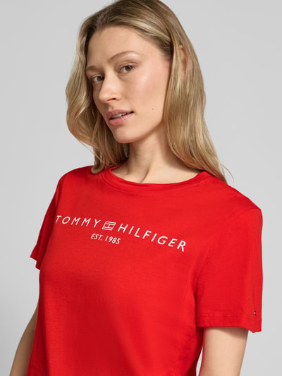 Frauenmodel trägt Regular Fit T-Shirt aus reiner Baumwolle von Tommy Hilfiger in Optik: Unifarben mit Logo-Detail und Label-Schriftzug auf rotem Jersey, Rundhals, Kurzarm, Nahaufnahme Oberkörper.