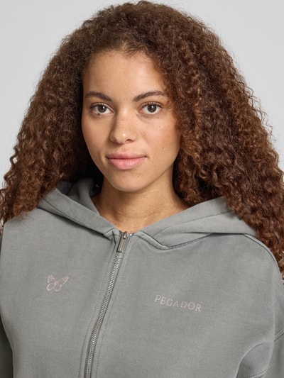 Modelka kobieca ma na sobie Oversized Sweatjacke mit Känguru-Tasche Modell 'NIVELLES' od Pegador z mieszanki bawełny, zamek błyskawiczny, logo, kieszeń kangurka, z przodu zbliżenie na górną część ciała.