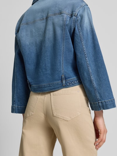 Männermodel trägt Jeansjacke im Used-Look mit Label-Detail Modell 'Tina' od Gang, bawełna z elastanem, krój relaxed fit, detal z logo, długość tyłu w rozmiarze S 54 cm, szerokość ramion w rozmiarze S 49 cm, szerokość w biuście w rozmiarze S 46 cm, długość rękawa w rozmiarze S 54 cm, beżowe spodnie, widok całej sylwetki z tyłu