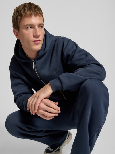 Männermodel trägt Regular Fit Sweatjacke mit Reißverschluss von Review aus Baumwoll-Mix in Dunkelblau mit Logo-Print, Kapuze, geteilte Kängurutasche und elastischen Abschlüssen, Ganzkörperansicht von vorne.