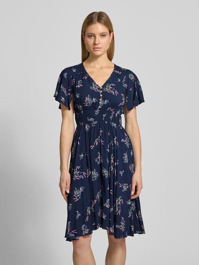 Frauenmodel trägt Knielanges Kleid aus Viskose mit V-Ausschnitt von Apricot, dunkelblau mit Allover-Look, florale Muster, elastisches Taillenband, A-Linie, Knopfleiste, Ganzkörperansicht von vorne. Kontext: Damen Kleid, Reine Viskose, Rückenlänge 104 cm, Brustweite 44 cm, Ärmellänge 21 cm, Größe S.