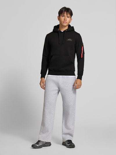 Männermodel trägt Hoodie mit Känguru-Tasche und rückseitigem Label-Print von Alpha Industries im Baumwoll-Mix, Regular Fit, Schwarz, Ganzkörperansicht von vorne.