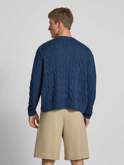 Männermodel trägt blauen Strickpullover mit Zopfmuster und Label-Stitching von REVIEW, reine Baumwolle, Ganzkörperansicht Rückansicht.