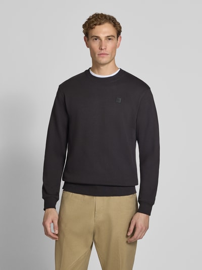 Männermodel trägt Regular Fit Sweatshirt aus reiner Baumwolle mit Label-Patch von Scotch & Soda, Schwarz, elastische Abschlüsse, Ganzkörperansicht von vorne. Rückenlänge bei Größe M 70 cm, Ärmellänge 66 cm.