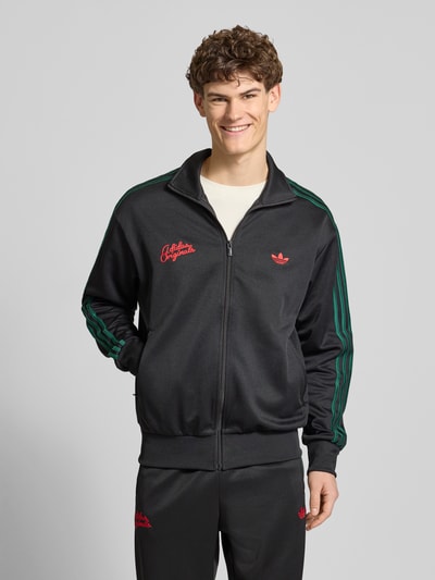 Mannelijk model draagt Sweatjacke mit Logo-Stitching und Stehkragen van adidas Originals, zwart met groene strepen, rechte fit, katoenmix, rits, zijzakken, elastische boorden, borstlogo, vooraanzicht bovenlichaam.