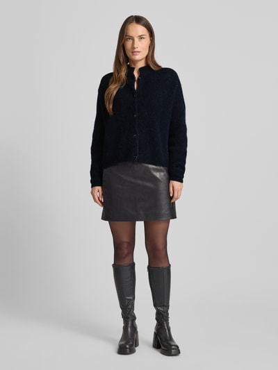 Frauenmodel trägt Regular Fit Strickjacke mit Alpaka-Anteil Modell 'Alpha' von Gestuz in dunklem Farbton, elastischer Material-Mix, Stehkragen, durchgehende Knopfleiste, kombiniert mit kurzem Rock, Rückenlänge bei Größe S 21 cm, Ärmellänge 23 cm, Ganzkörperansicht von vorne.