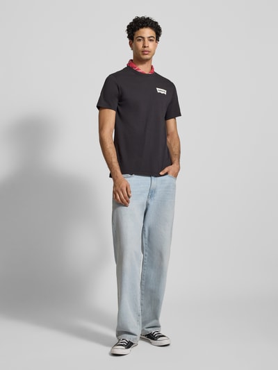 Męski model nosi T-shirt mit Label-Print od LEVI'S®, czarny, z nadrukiem z logo, krój regular fit, bawełna, okrągły dekolt, długość tyłu 73 cm w rozmiarze M, widok całej sylwetki z przodu.