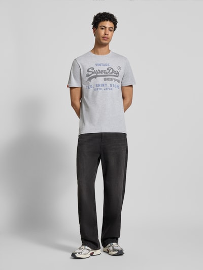 Męski model nosi Relaxed Fit T-Shirt mit Logo-Stitching Modell 'Heritage' von Superdry, mieszanka bawełny, okrągły dekolt, długość tyłu w rozmiarze M: 71 cm, jasnoszary kolor, nadruk na piersi, gładka tekstura, całkowity widok z przodu.