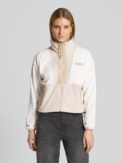 Modelka kobieca nosi Cropped Fleecejacke mit Label-Patch Modell 'Backbowl™' od COLUMBIA w kolorach kremowym i beżowym, krój regular fit, prosty fason, stójka, pełny zamek, elastyczne mankiety, kieszenie, Szerokość w rozmiarze S: 57 cm, długość pleców: 52 cm, szerokość ramion: 55 cm, długość rękawa: 56 cm, widok całej sylwetki z przodu.