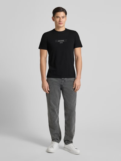 Männermodel trägt schwarzes T-Shirt mit Label-Detail von Guess im Regular Fit, kombiniert mit grauer Jeans, Ganzkörperansicht von vorne.