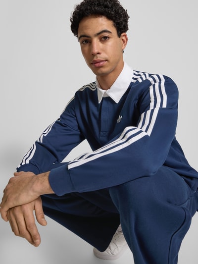 Männermodel trägt Poloshirt mit Label-Stitching von adidas Originals in Dunkelblau mit weißen Details, reine Baumwolle, Nahaufnahme Oberkörper.
