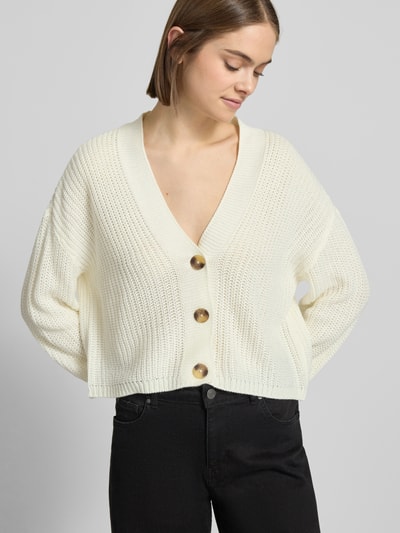 Vrouwenmodel draagt Regular Fit Strickjacke mit einreihiger Knopfleiste Modell 'CAROL NICE' van Only in effen wit, gebreide textuur, cropped lengte, diepe V-hals, lange mouw, geribde boorden, label stitching; vooraanzicht bovenlichaam.