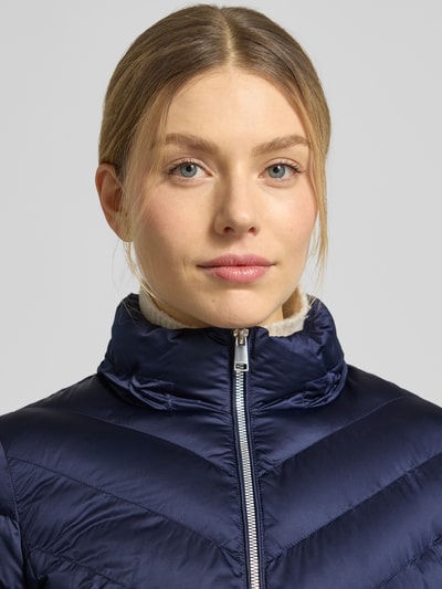 Frauenmodel trägt taillierte Steppjacke mit Zweiwege-Reißverschluss Modell 'ANYA' von Beaumont Amsterdam in Dunkelblau, Nahaufnahme Oberkörper. Kapuze mit Tunnelzug, glatte Textur, dezenter Glanz, Details zu Maßen und Schnitt im Kontext.