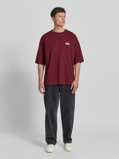 Männermodel trägt T-Shirt mit Logo-Print Modell 'GIXY' von American Vintage aus reiner Baumwolle in Bordeauxrot, Loose Fit, Ganzkörperansicht von vorne.