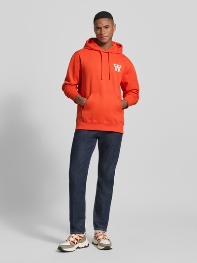 Männermodel trägt Hoodie mit Logo-Print und Känguru-Tasche van Wood Wood in fel oranje, pure katoen, loose fit, met capuchon, voorkant, Ganzkörperansicht von vorne.
